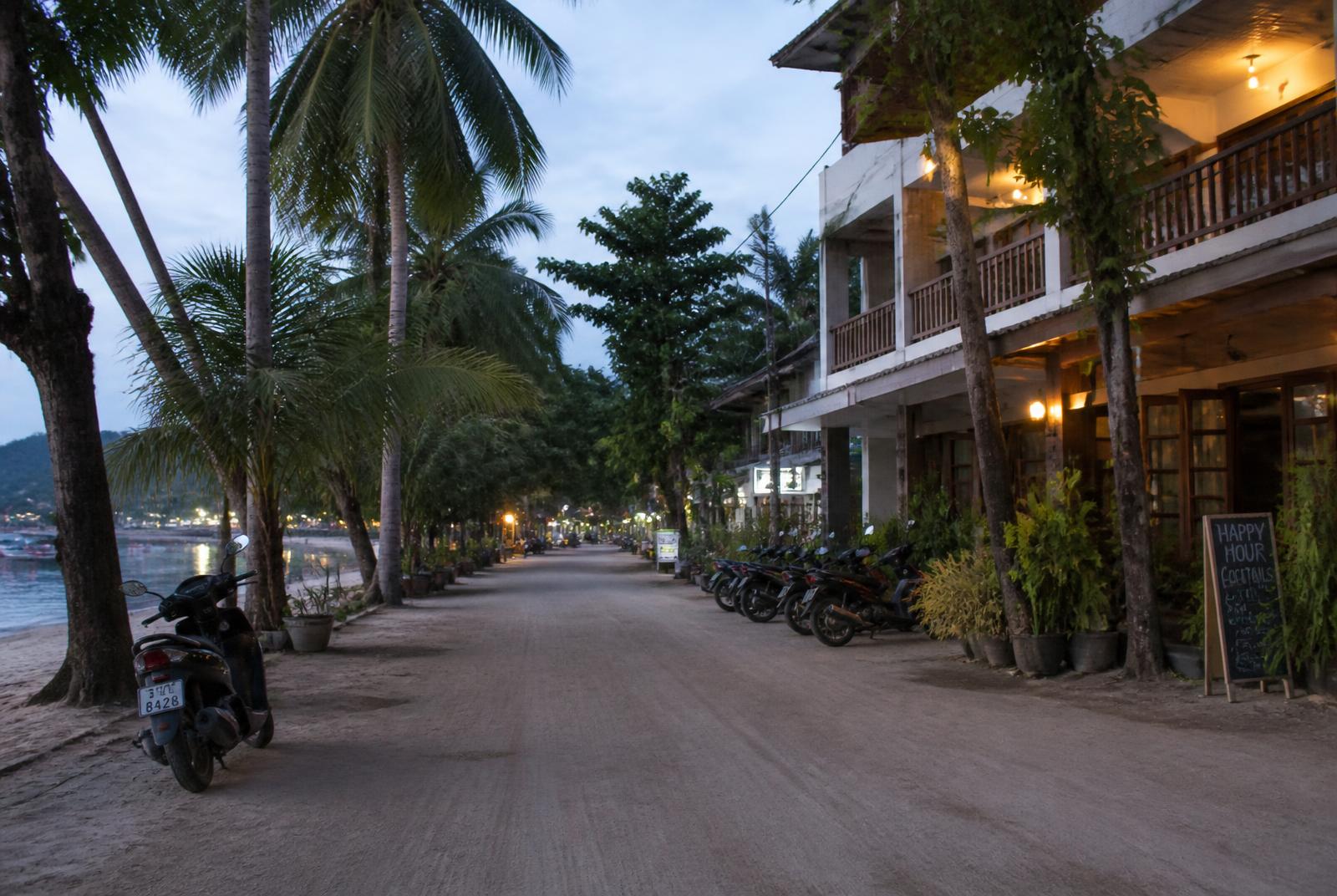 Quiet Thai island beachfront street at dusk lined with low-rise guesthouses and small cafes